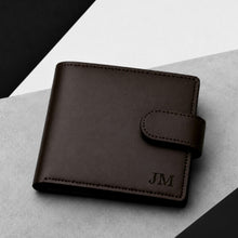 Load image into Gallery viewer, A dark brown leather bifold wallet with a snap tab closure and debossed initials 'JM' on the front, shown on a light gray textured surface with black geometric background shapes.