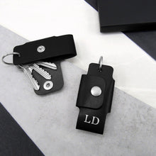 Load image into Gallery viewer, A handmade black personalised leather key case on a grey and white stone surface surrounded by black gift packaging for him.