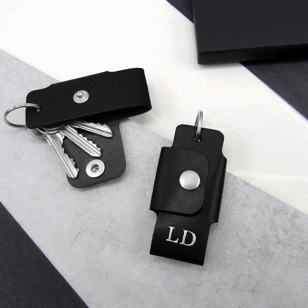A handmade black personalised leather key case on a grey and white stone surface surrounded by black gift packaging for him.