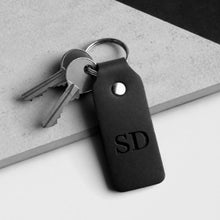 Load image into Gallery viewer, A black leather keychain with an debossed 'SD' monogram and attached keys, photographed on a light grey surface with a dark corner in the background.