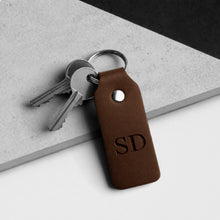 Load image into Gallery viewer, A dark brown leather keychain with a metal key ring holding two keys, featuring the debossed initials 'SD' on a clean, diagonally split grey and white background.