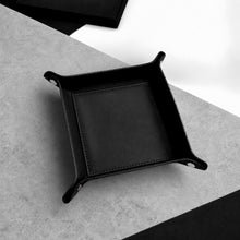 Load image into Gallery viewer, A Handmade Black Leather Desk Tidy Valet Tray with raised corners and polished silver snap fasteners, photographed on a light grey textured stone surface with a black and white geometric background