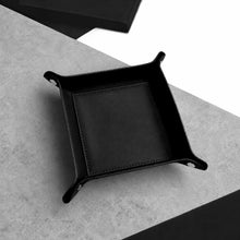Load image into Gallery viewer, A black Handmade Leather Desk Tidy Valet Tray with raised corners and stitched edges, photographed on a light grey textured surface with a dark box partially visible in the background.