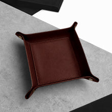 Load image into Gallery viewer, A square dark brown Handmade Leather Desk Tidy Valet Tray with raised corners and brass-tone snap closures, photographed on a grey textured surface with a black box partially visible in the background.
